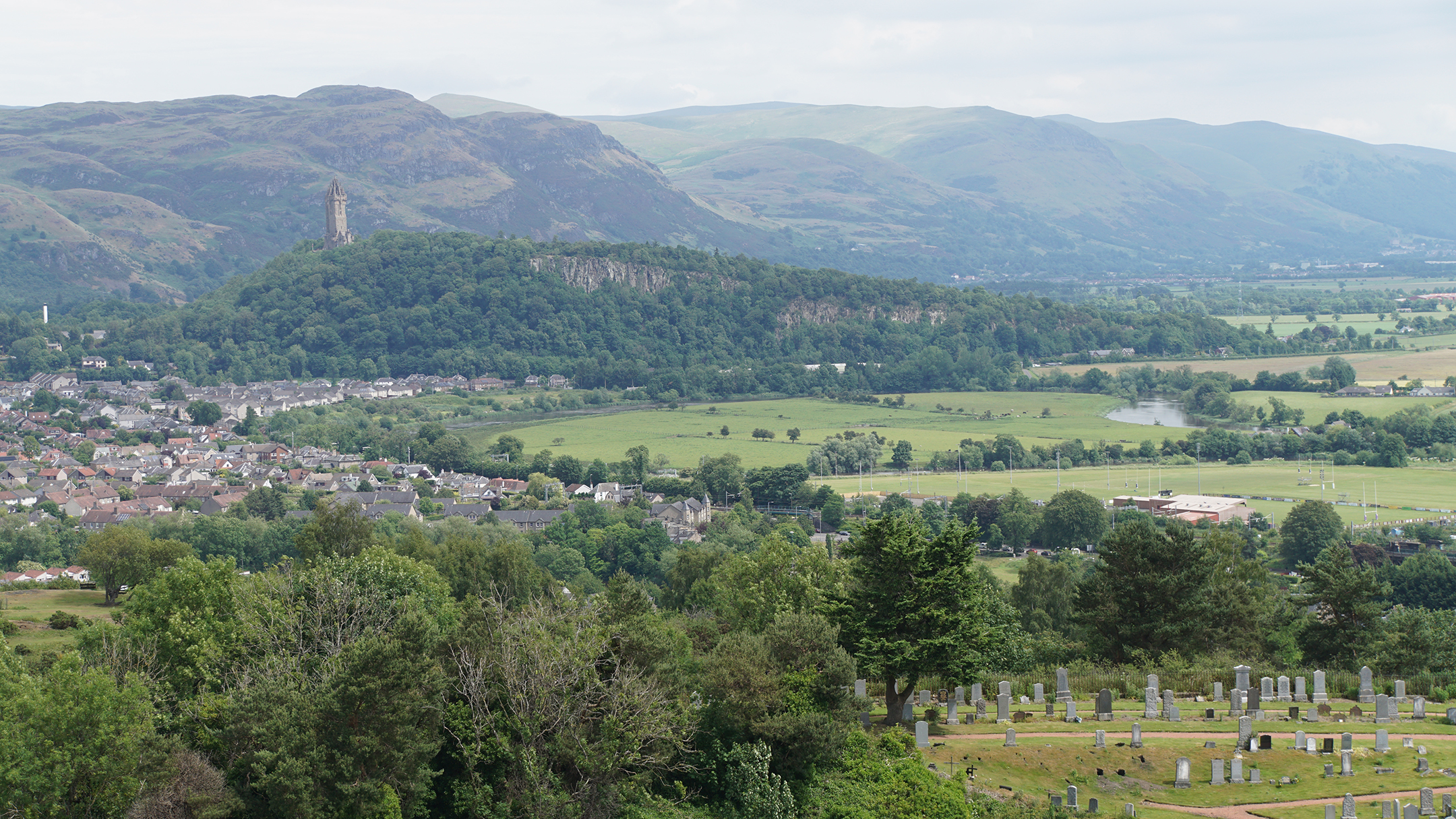 A tree covered hill with a stone tower overlooks a field with a river running along side
