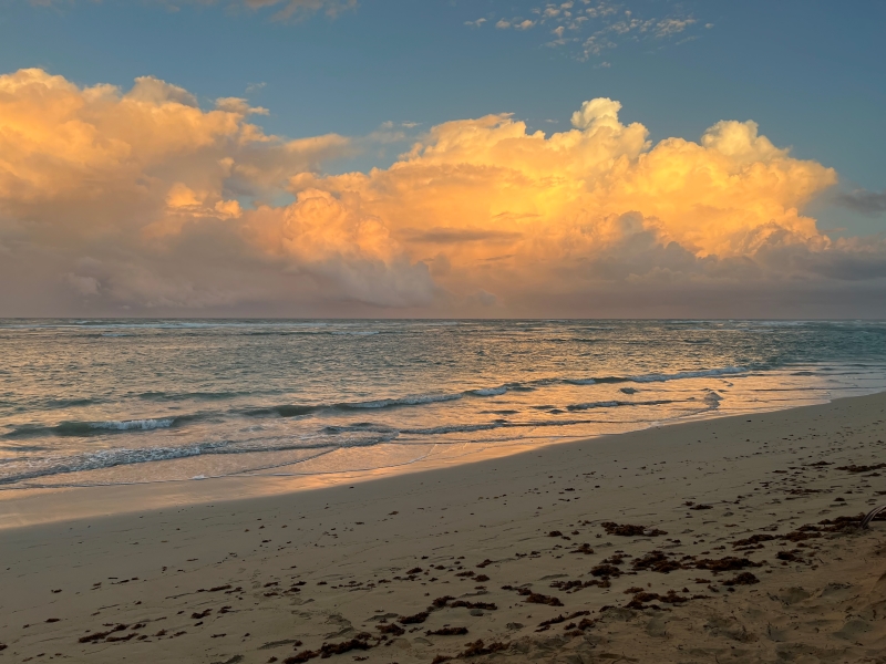 Puffy clouds reflect the oranges and reds of the setting sun above waves softly lapping at a sandy beach