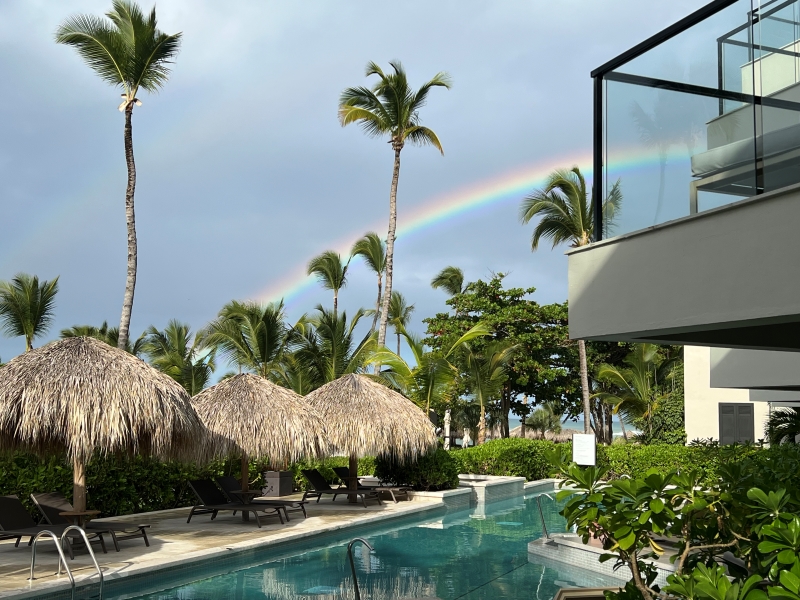 A rainbow cuts across the sky beyond palm trees and thatched shade structures near a pool