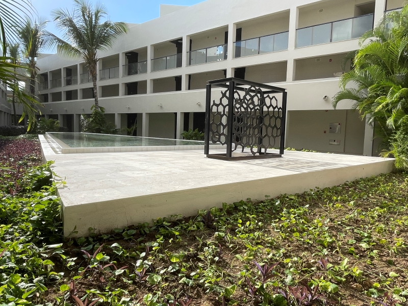 A sculpture sits on a tile patio next to a reflecting pool surrounded by plants in a courtyard lined by a three-story building