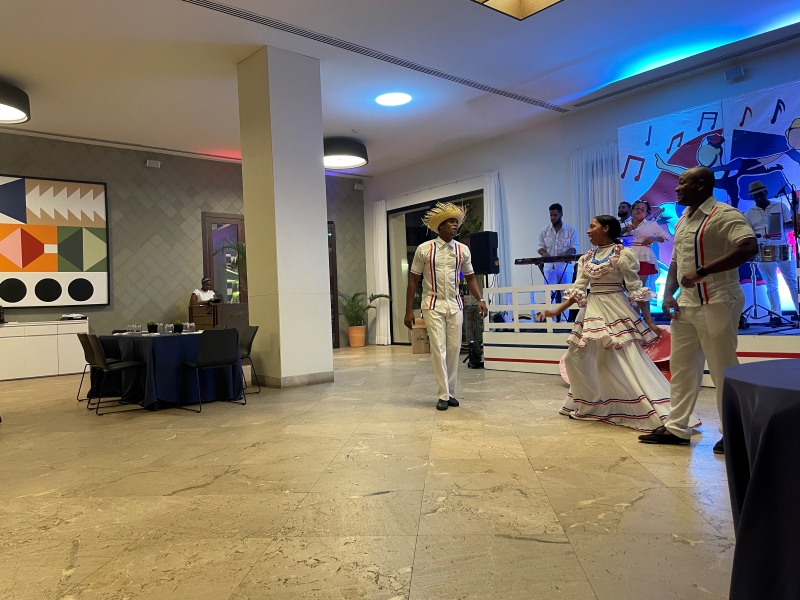 Men and a woman in white clothes with red and blue trim dance while a band plays behind them