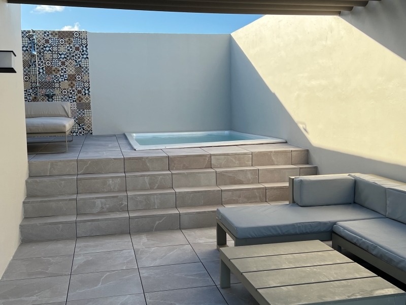 Tan walls surround a plunge pool, two-person outdoor bed, a shower, and seating area open to the blue sky above