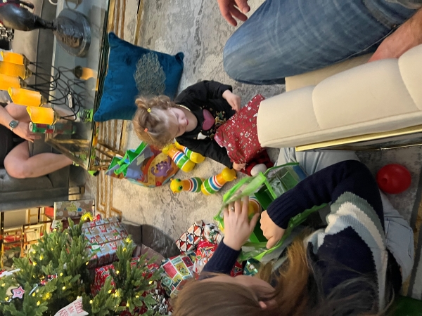A young girl tears wrapping paper off a boxed gift near the Christmas tree while her family looks on
