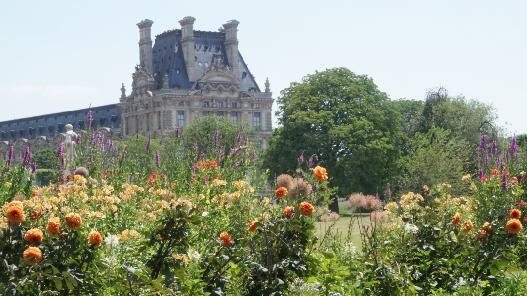 Orange, yellow, and purple flowers bloom with trees and an ornate building with a slate roof beyond
