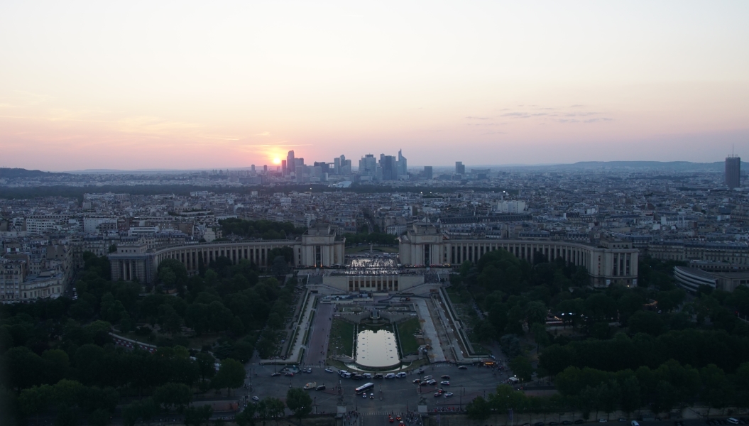 The sun sets behind a distant skyline with lower buildings in the foreground around a park like plaza