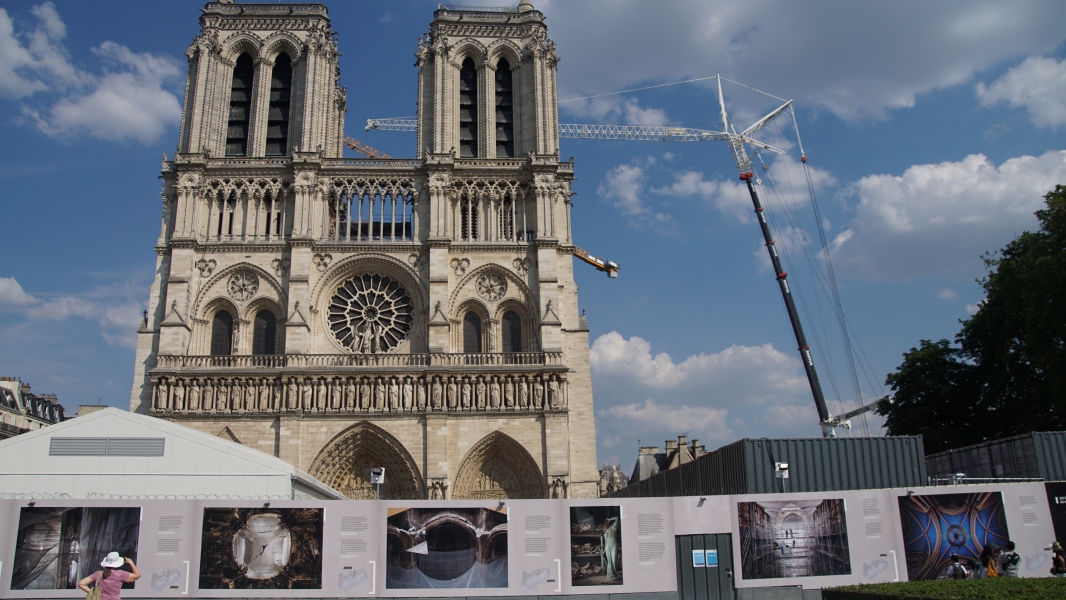 A construction wall with photos of a fire damaged building stand in front of a gothic cathedral with a crane visible to one side