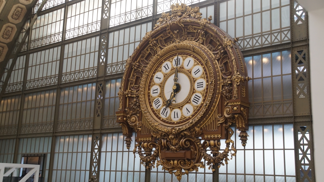 An ornately detailed gold trimmed clock hangs on a wall with steel framing and translucent glass