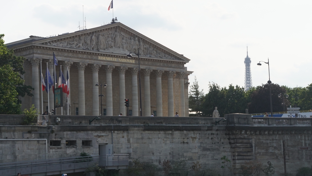 A building with a stone façade and Grecian details stands near the river with the Eiffel Tower visible in the distance