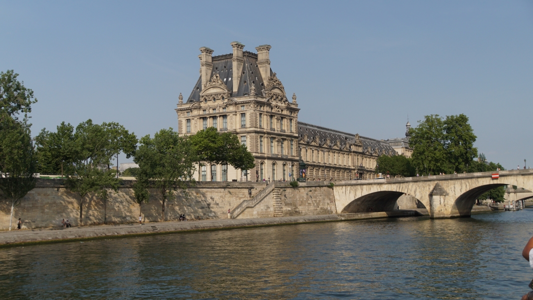 One three story stone building with ornate details and a high mansard roof next to a wall along a river