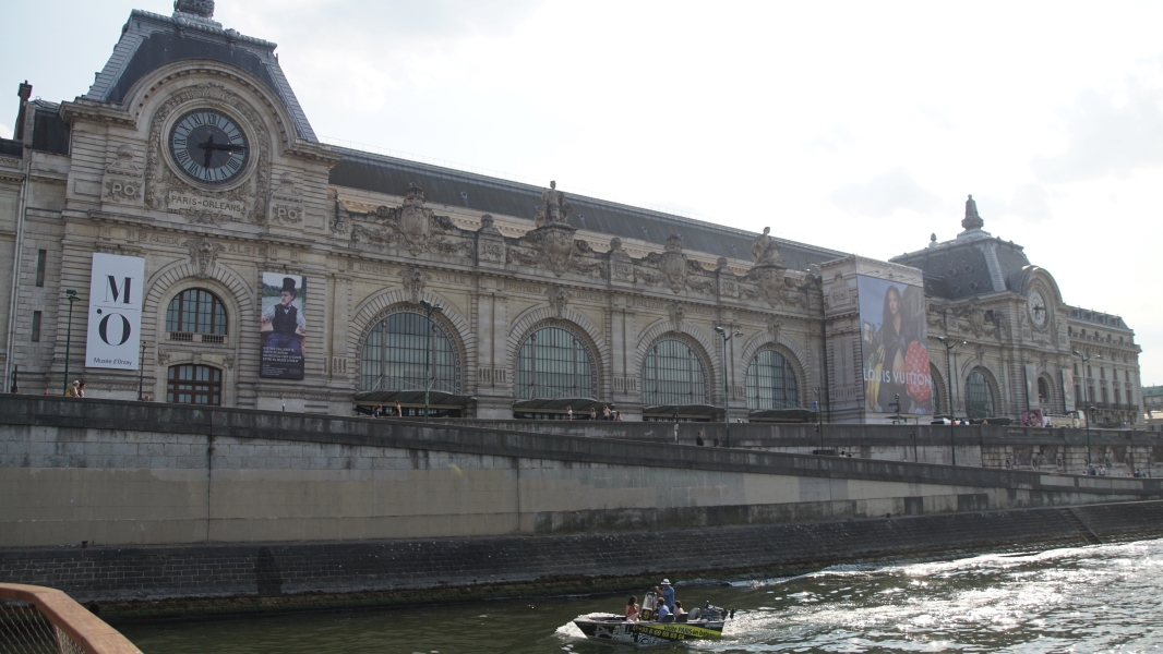 A stone building with large arched openings infilled with steel frames and glass and two entry elements with clocks along a river