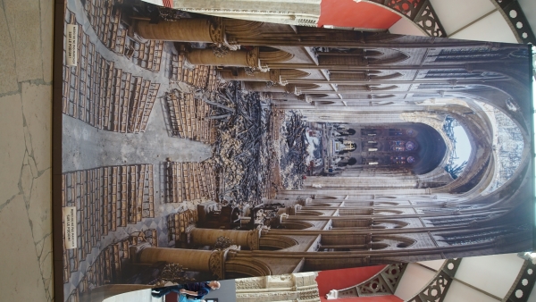 A banner hangs from the ceiling of a large space showing the inside of a tall cathedral space with light streaming through fire damaged holes in the roof