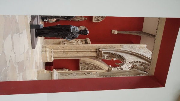 A tall dark statue stands amongst ornately carved stone elements inside a museum gallery