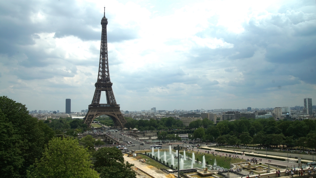 A metal tower extends into the sky with a bridge and park w/ fountains in the foreground