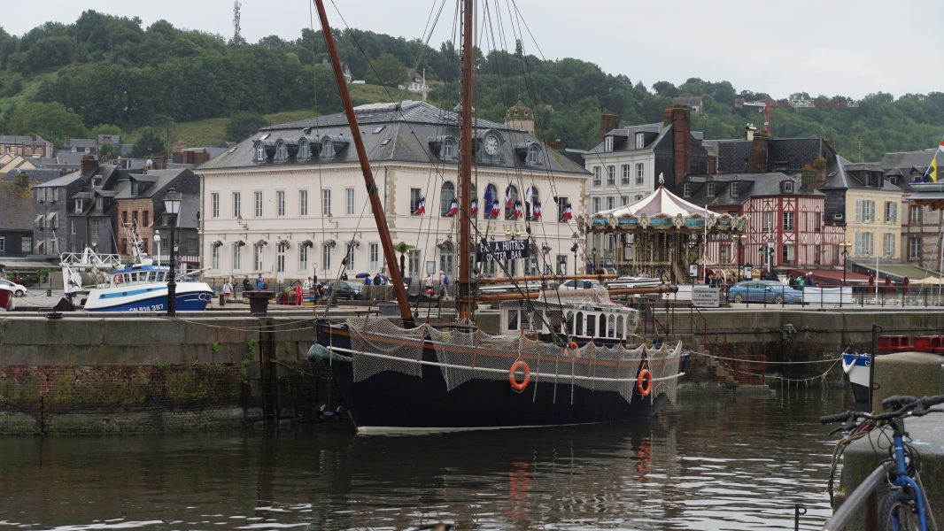 A black hulled ship with tall masts and fish nets sits against a stone pier near tall buildings along the waterfront