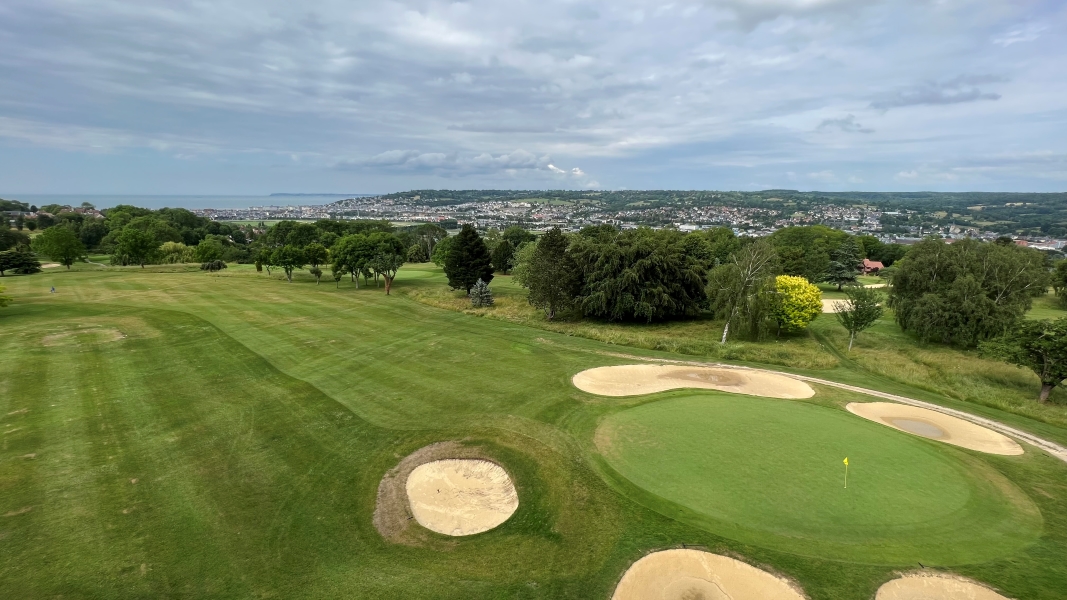 The green of a golf course is surrounded by sound traps looking out over a valley with the ocean visible in the distance