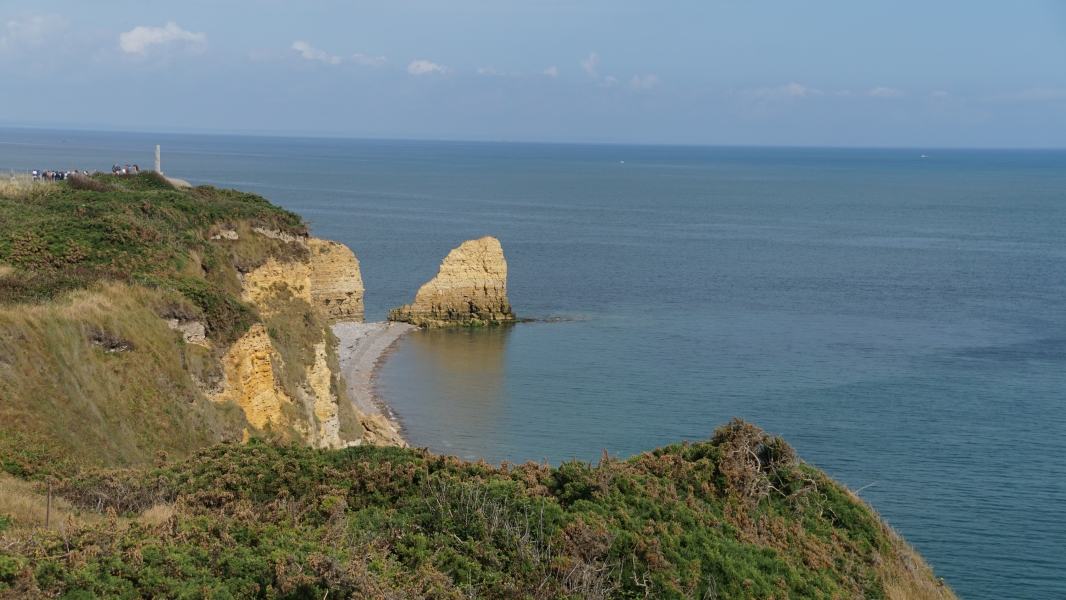 Tan cliffs with scrubby green tops extend above a narrow rocky beach with ocean extending beyond the horizon