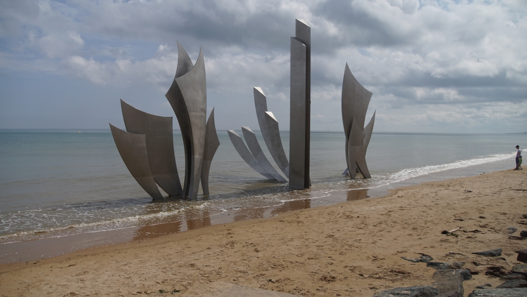 Curved metal forms extend out of shallow water on a sandy beach