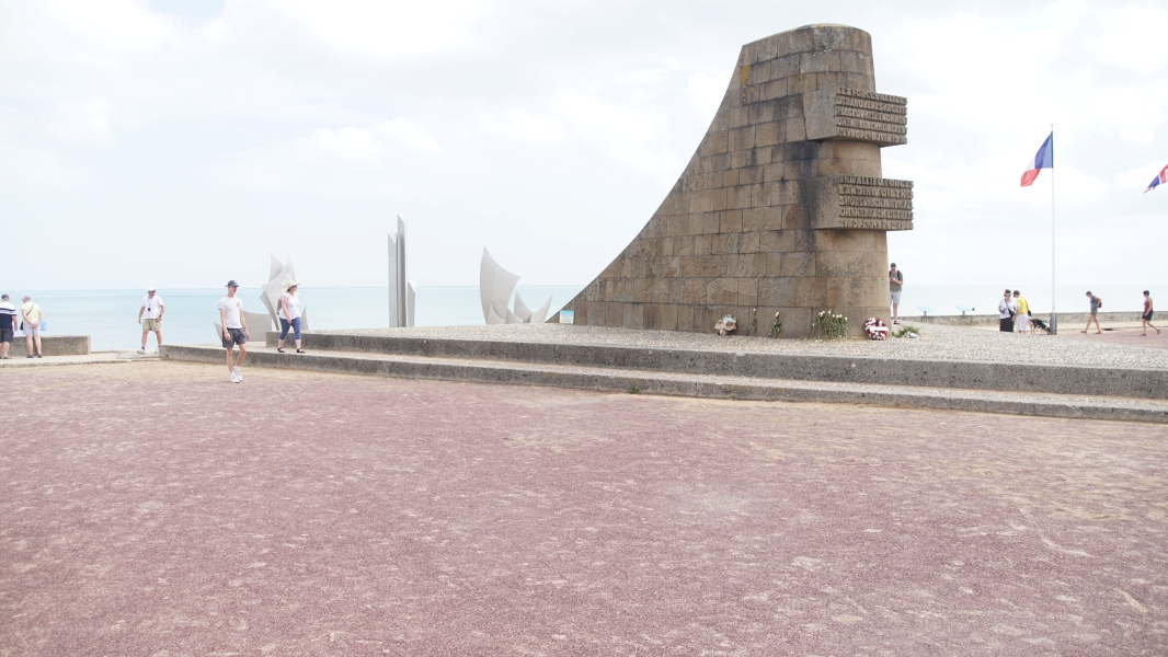A stone monument with a curved top appears to be coming out of the beach with flags from various countries flying nearby