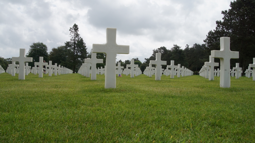 White stone crosses in rows stick out of the green grass extending into the distant trees under cloudy skies