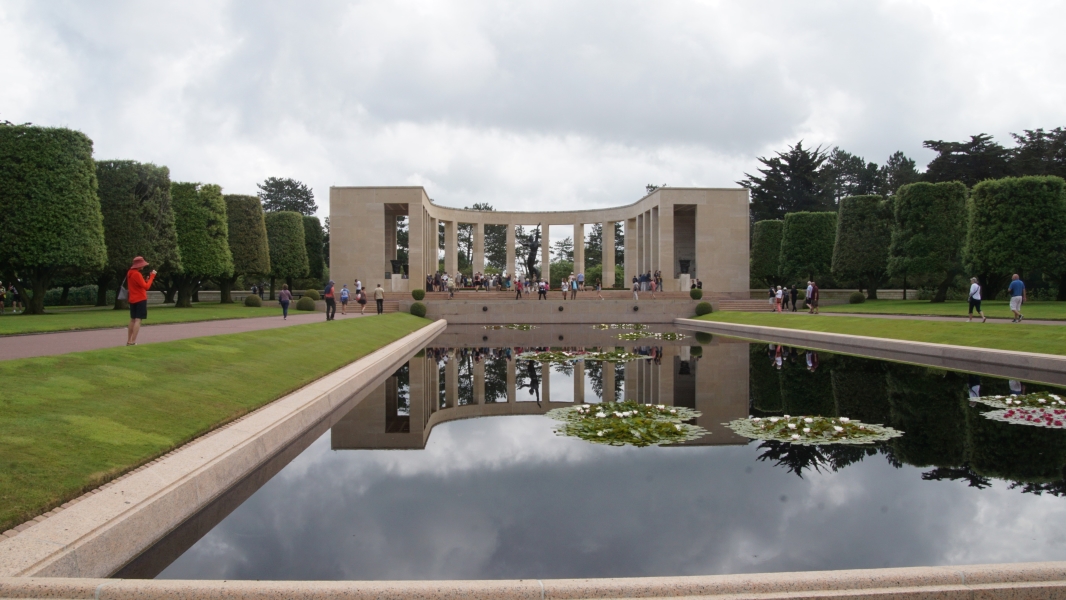 A tan stone memorial structure surrounds a sculpture at the far end of a reflecting pool