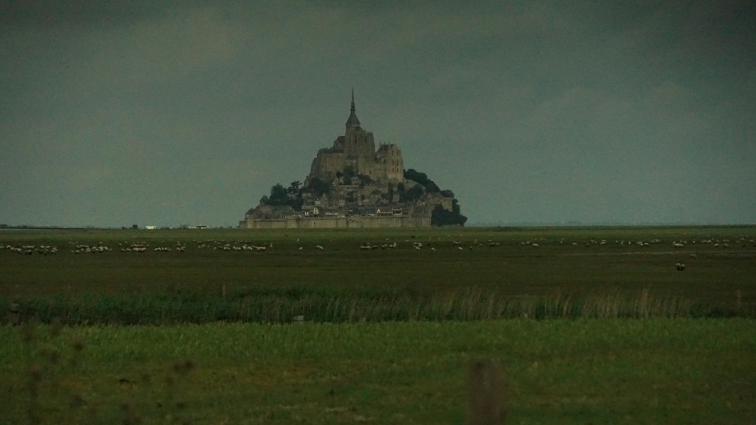 A rock mount with a tall church-like structure at its crown extends above the flat grassland dotted with grazing sheep