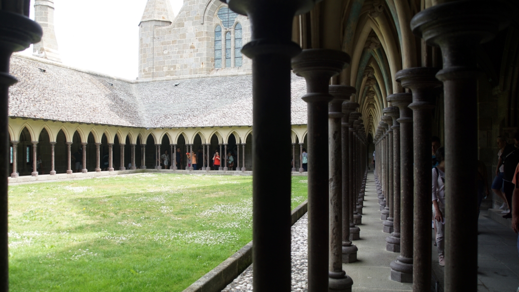 Rows of narrow columns with arched connections extend around a grass area connect to the stone wall of a church