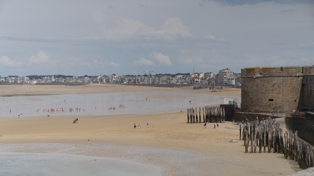 A thin sheen of water on a large sandy expanse of beach leads to the stone wall of a medieval keep with modern buildings lining the beach in the distance