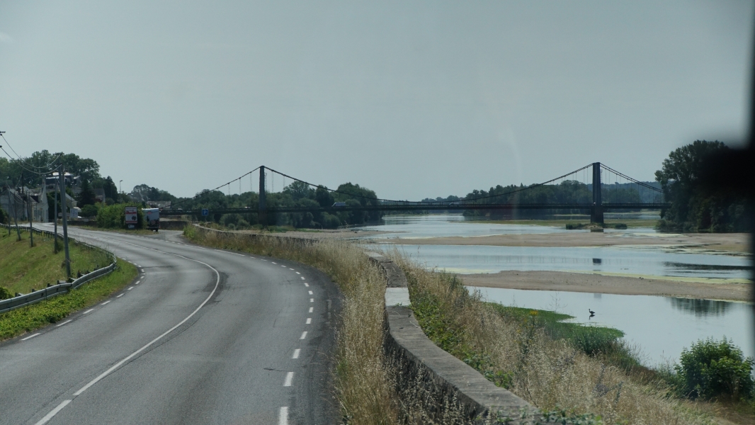 A road curves along the bank of a shallow river with several sandbars and a cable suspension bridge