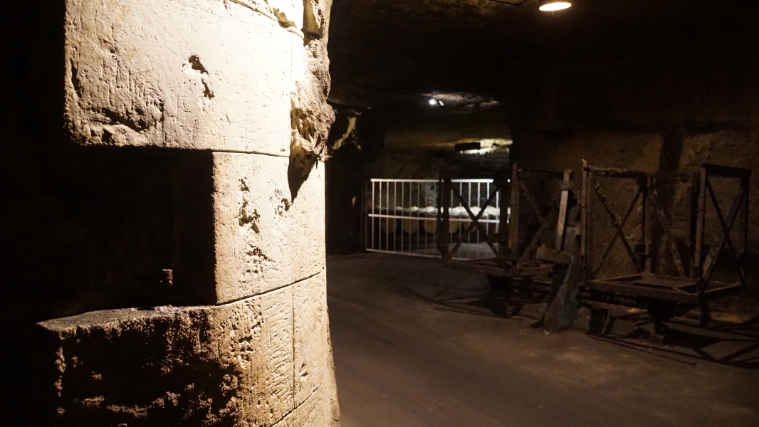 A carved limestone wall in a dark space with barrels used for aging wine and a device used to turn and store sparkling wine bottles visible beyond