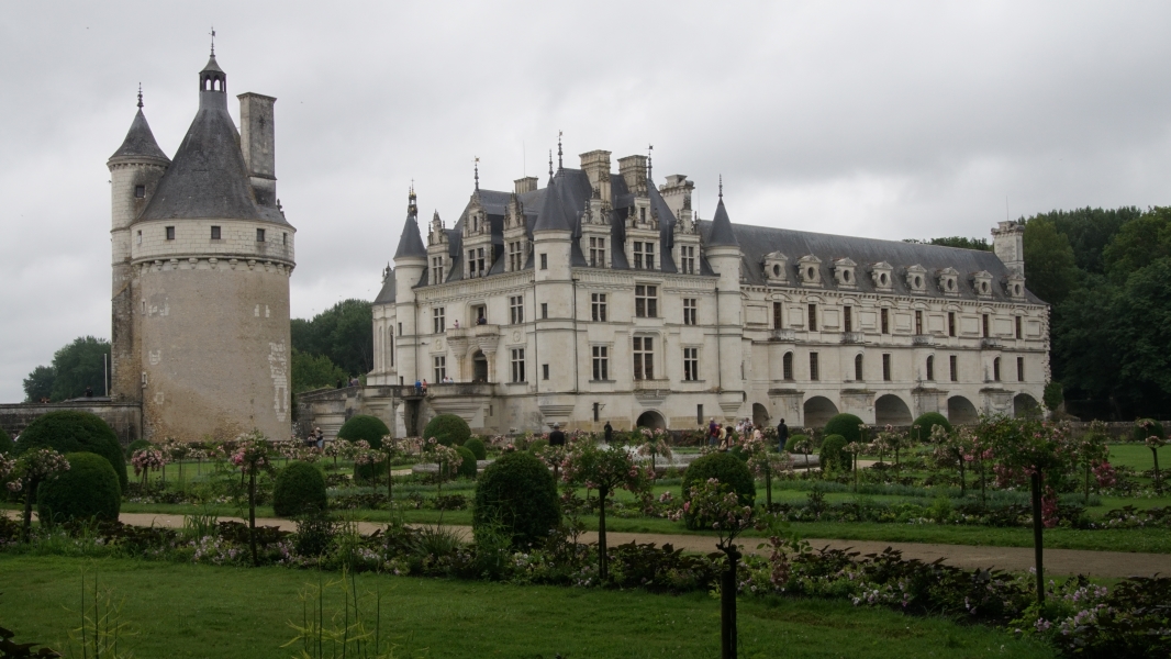 A medieval tower and white stone chateau beyond a garden with stone paths and short blooming trees