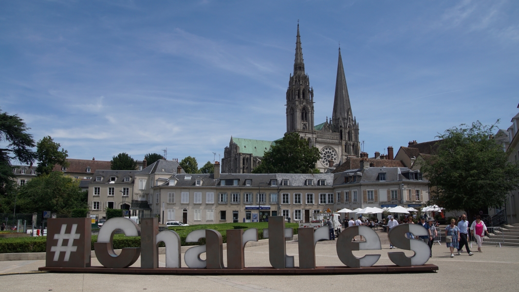 Sculpture letters five-foot high spell #Chartes with two bell towers of a church extend above several buildings in the distance