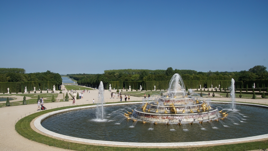 A large circular fountain with three tiers of gilded statues stands at the end of a long path between tall, maintained trees
