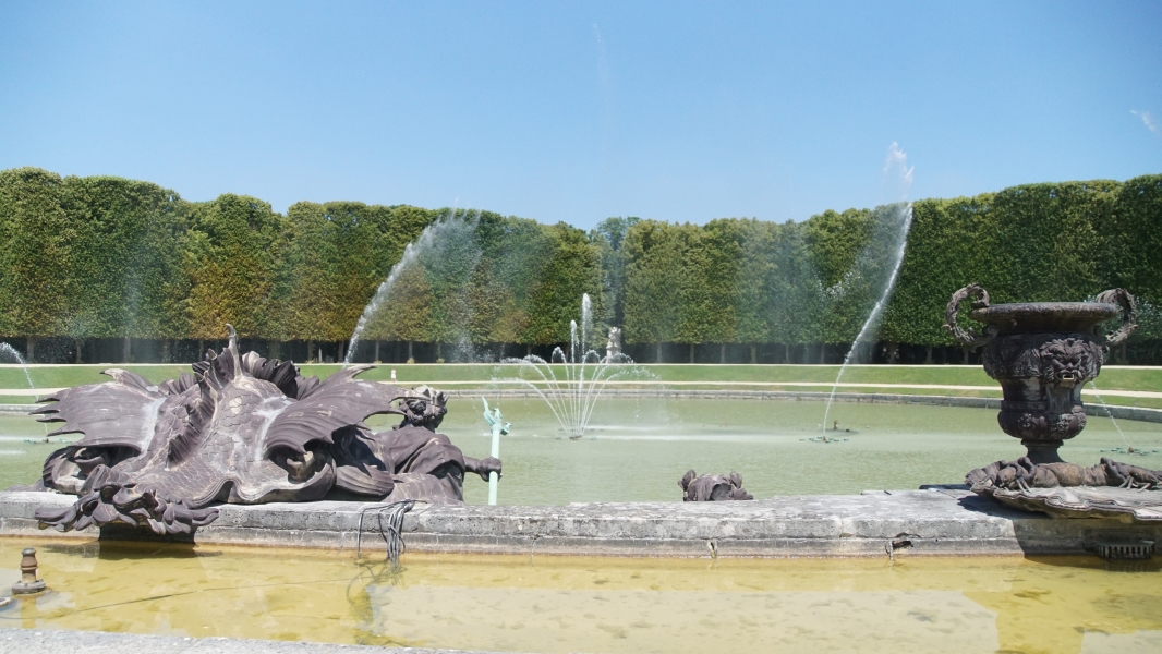 Square cut trees line the far side of a pool of water with fountains shooting water into the air in front of a statue of Poseidon