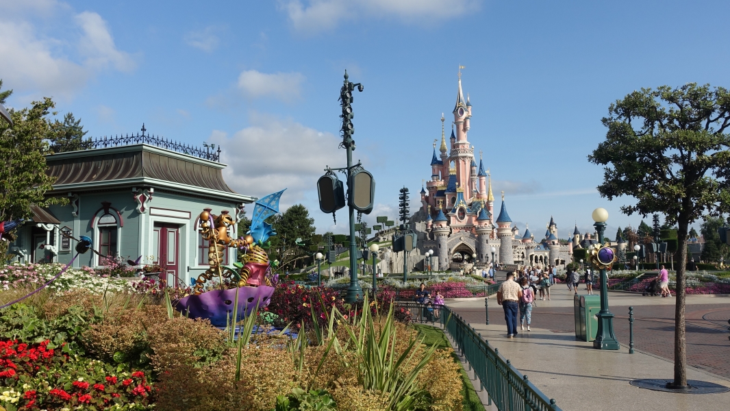 A castle with pink and gray walls rises beyond walkways and flower beds with a sculpture of Winnie the Pooh and Tigger in an umbrella