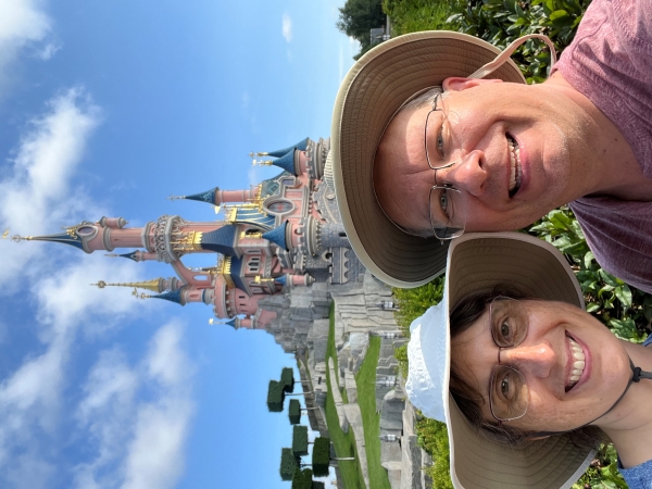 James and Julie in front of green hills leading up to a castle with pink walls and blue roofs