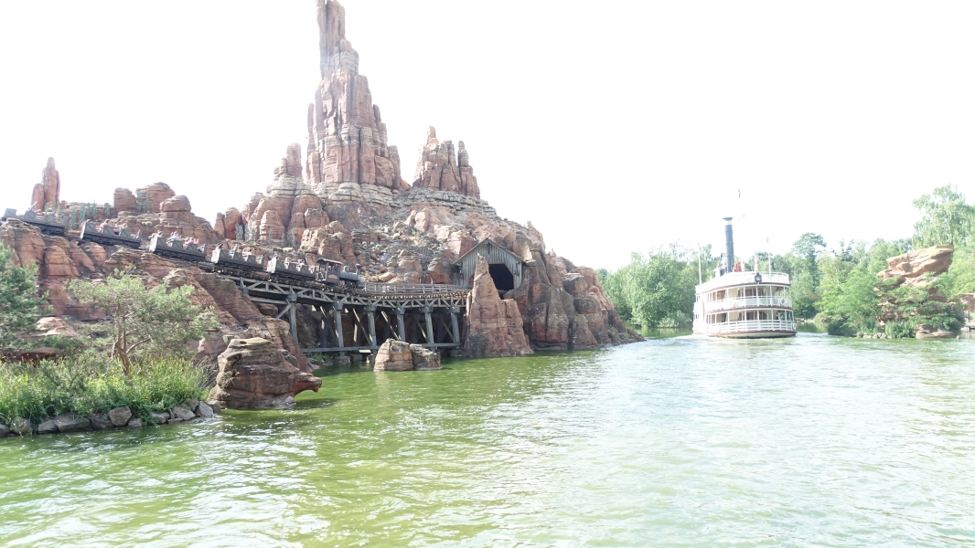 An old-style paddle boat moves through green water while a roller coaster train runs along tracks on an island of red stone buttes