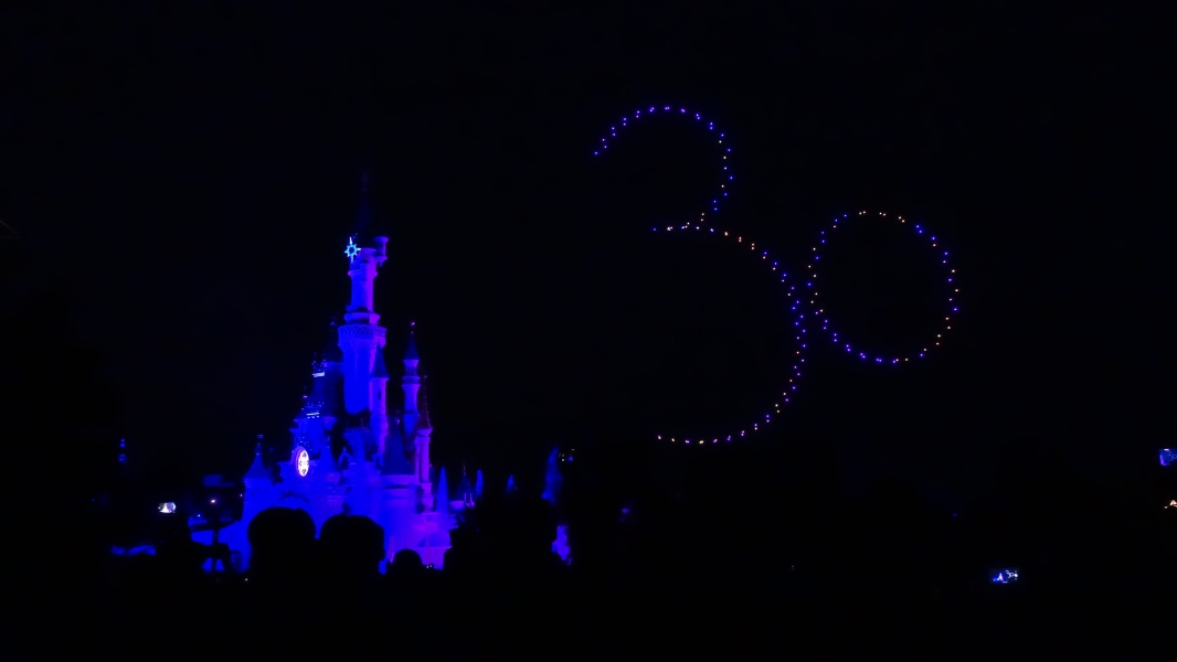 A dark purple castle in a dark night sky with drones forming a 30 in the sky