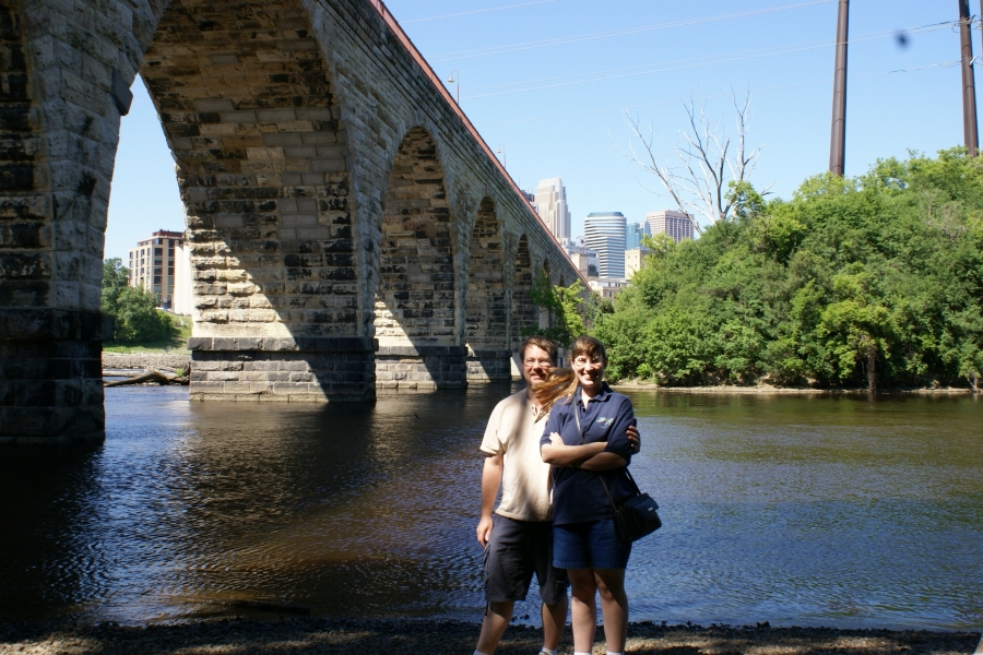 James and Julie stand next to a river with a stone arch bridge running to a downtown district with high rise buildings