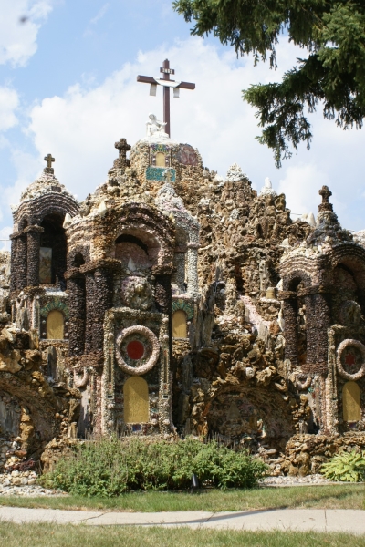 Shells and stones are stuck to concrete shapes forming a sort of temple facade with a cross at the highest point