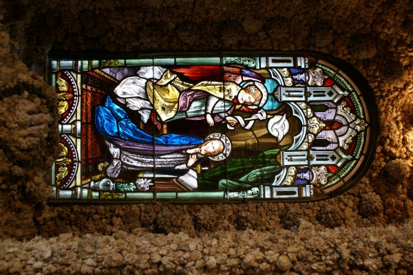 A stained-glass window showing a woman kneeling while an angel and dove visit her set in a rough stone wall