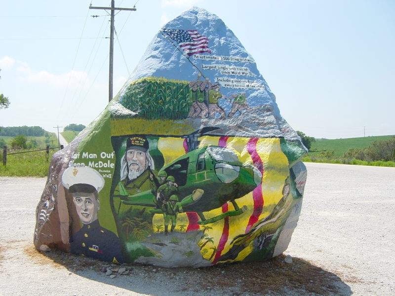 A painted image on a large stone shows several men raising a flag in front of a corn field, men exiting a helicopter onto a field with palm trees by a man with white hair and beard wearing a Vietnam Veteran cap