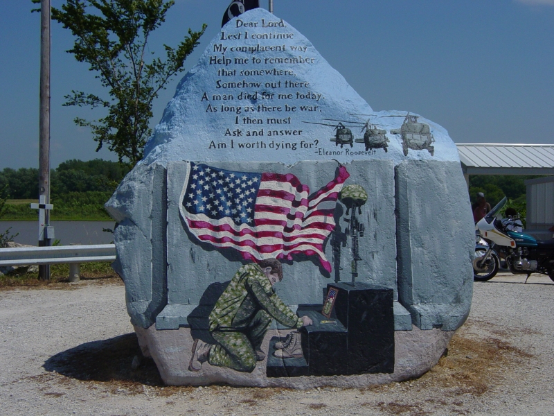 A painted image on a large stone shows a service person kneeling at a memorial below the words Dear Lord, Lest I continue My complacent way, help me to remember that somewhere, somehow out there a man died for me today. As long as there be war, I then must ask and answer Am I worth dying for? – Eleanor Roosevelt