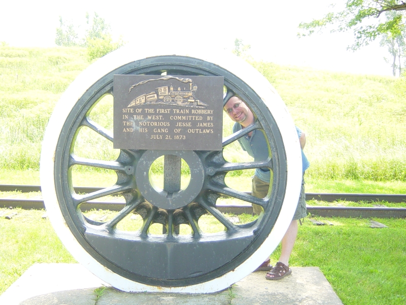 James looks through the spokes of a drive wheel from an old train with a sign reading Site of the first train robbery i nthe west. Committed by the notorious Jesse James and his gang of outlaws July 21, 1873.