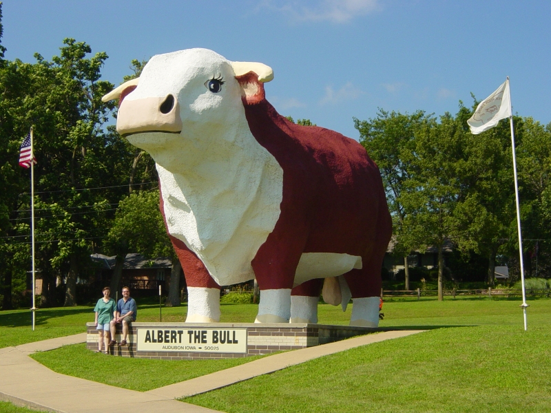 Julie and James sit on a pedistal next to a very large statue of a red body bull with a white head and hoofs