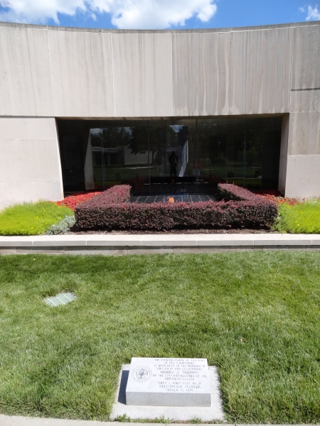 Red shrubs surround a black tile with a flame burning in front of a glass window with a statue of President Harry Truman looking out