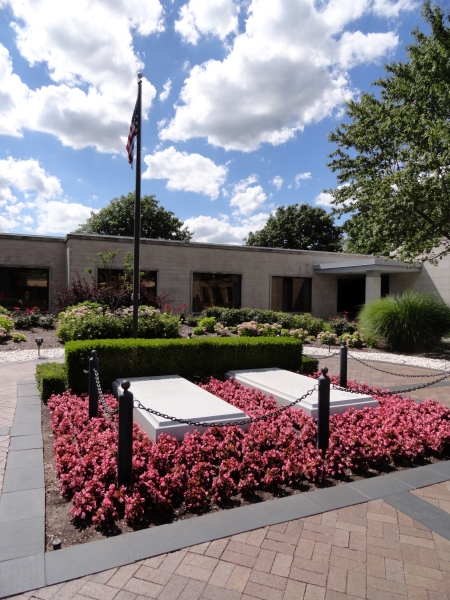 Pink blooming flowers surround two bright white marble crypts in a courtyard filled with other flowering plants and a flagpole
