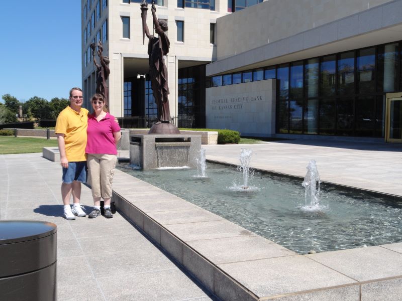 Julie and James stand near a fountain with a bronze sculpture of a woman holding a torch and banner on a staff outside a large building with marble and glass and a sign reading Federal Reserve Bank of Kansas City