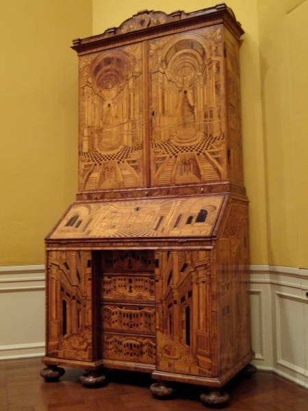 An intricately designed wood bureau with glossy inlays of different colored wood creating images of a checkered floored building with soring columns and arched ceilings
