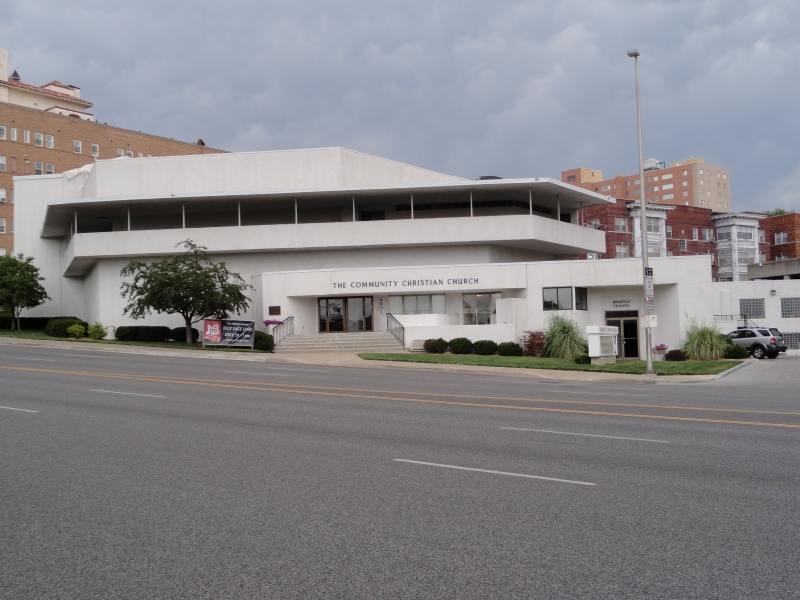 A stark white two-story building with prominent horizontal elements sets along a street surrounded by red brick structures