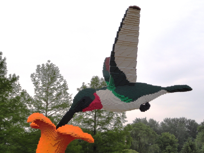 A very large, colorful LEGO hummingbird is supported by the beak which extends into an orange blossom
